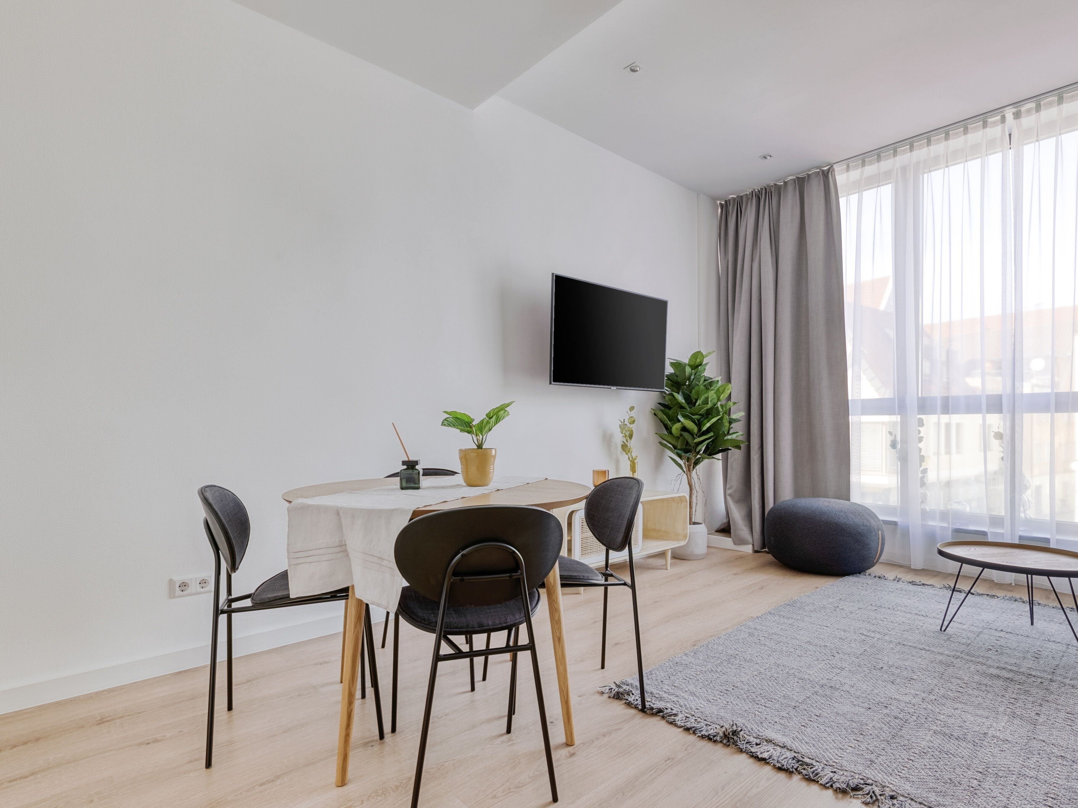En moderne stue med spiseplads, et fladskærms-TV og store vinduer med gardiner.