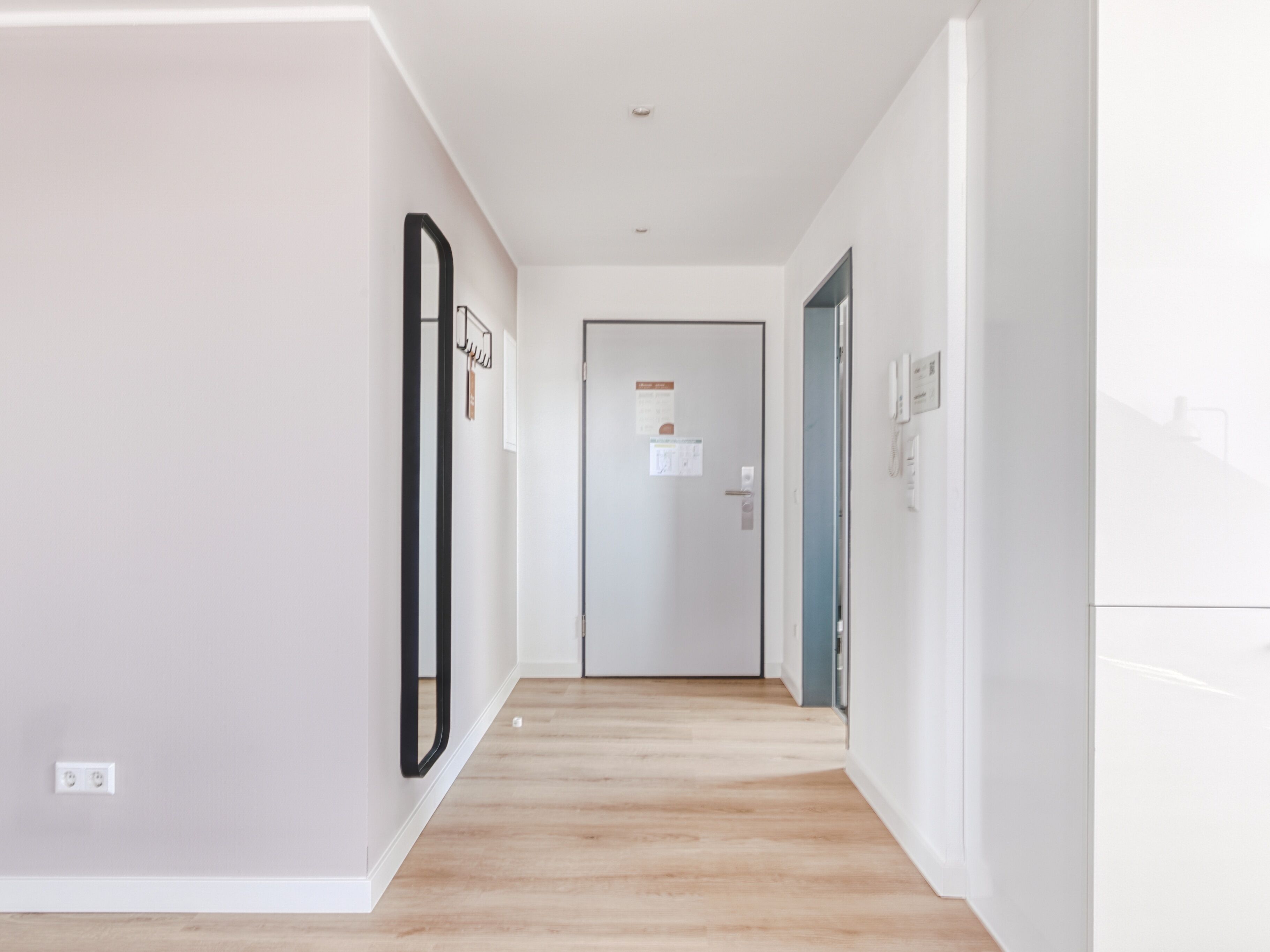 Un corridoio moderno e luminoso con pavimento in legno, pareti bianche e una porta in fondo.