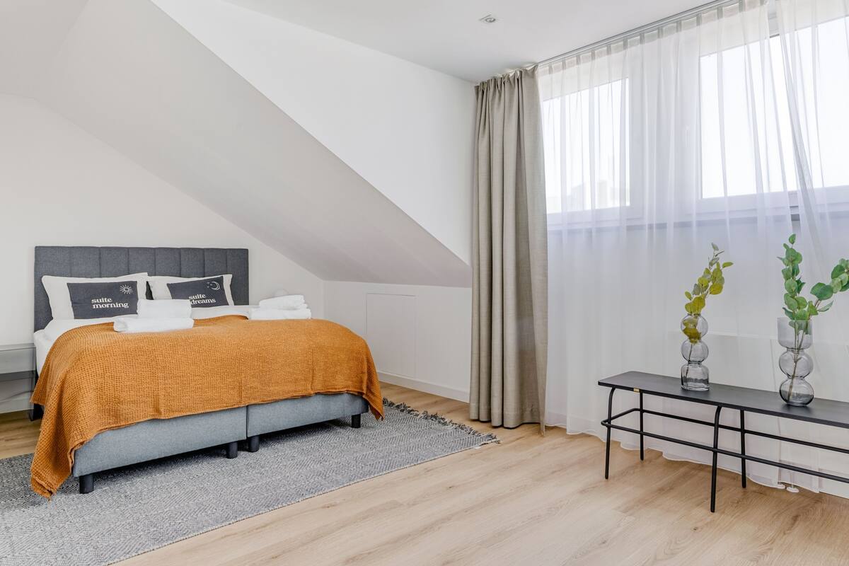 A modern bedroom with a sloped ceiling, a bed with a grey headboard, a mustard-colored blanket, and a small table with two glass vases.