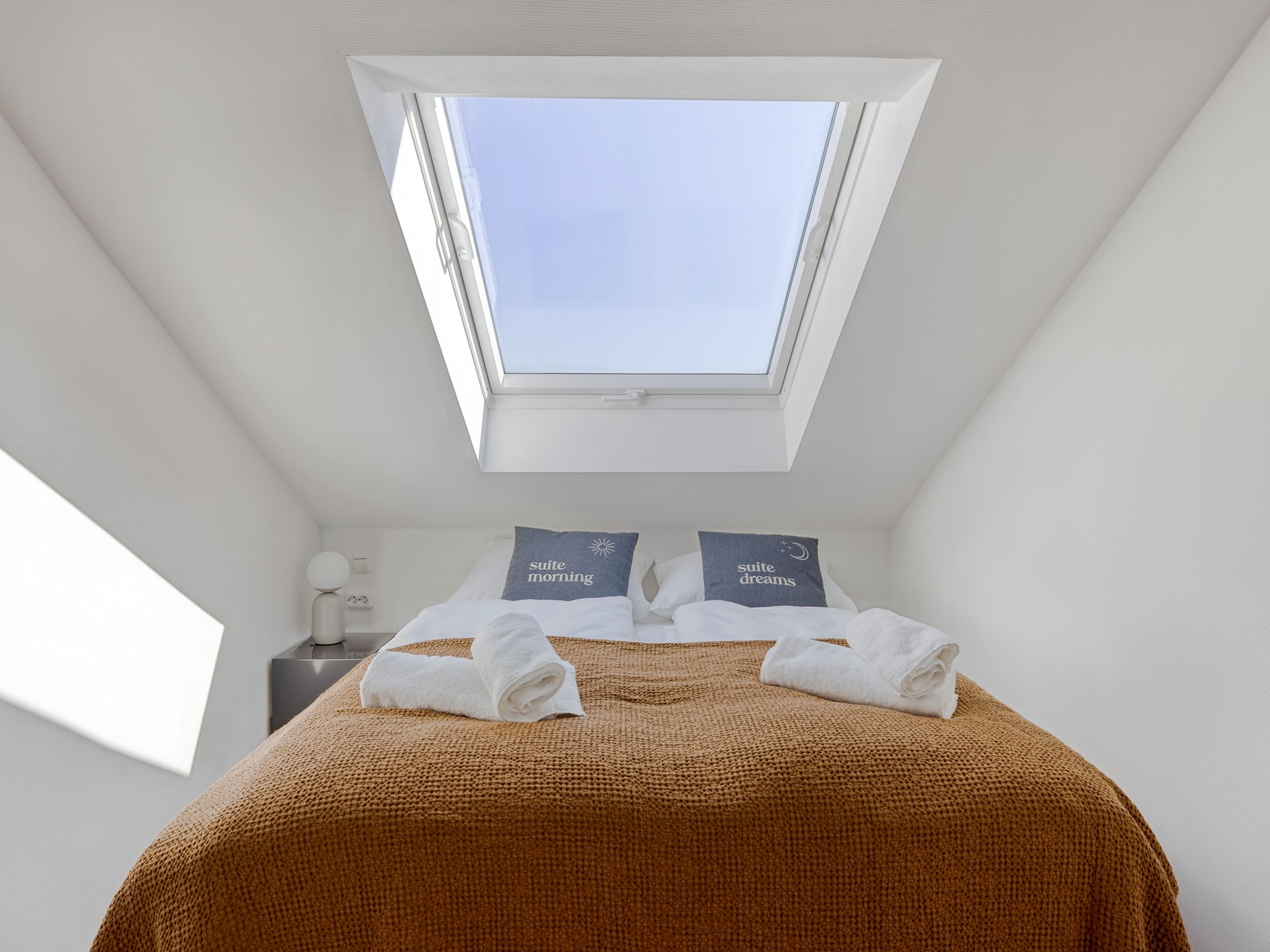 A bedroom with a skylight, a bed with two pillows, and a bedside table.