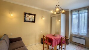 Un coin repas avec une table recouverte d’une nappe rose, un lustre, une œuvre d’art florale encadrée au mur et une fenêtre ornée de rideaux légers.