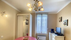 Une pièce compacte comprenant une table à manger, des chaises, un petit coin cuisine, un luminaire au plafond et une fenêtre avec des rideaux légers.