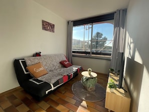 A cozy living room with a sofa, a small round table, and a window with a view of the outside. - Cozy 2-Room Apartment with Parking, Close to Beach and Center in Saint-Cyprien (Saint-Cyprien)