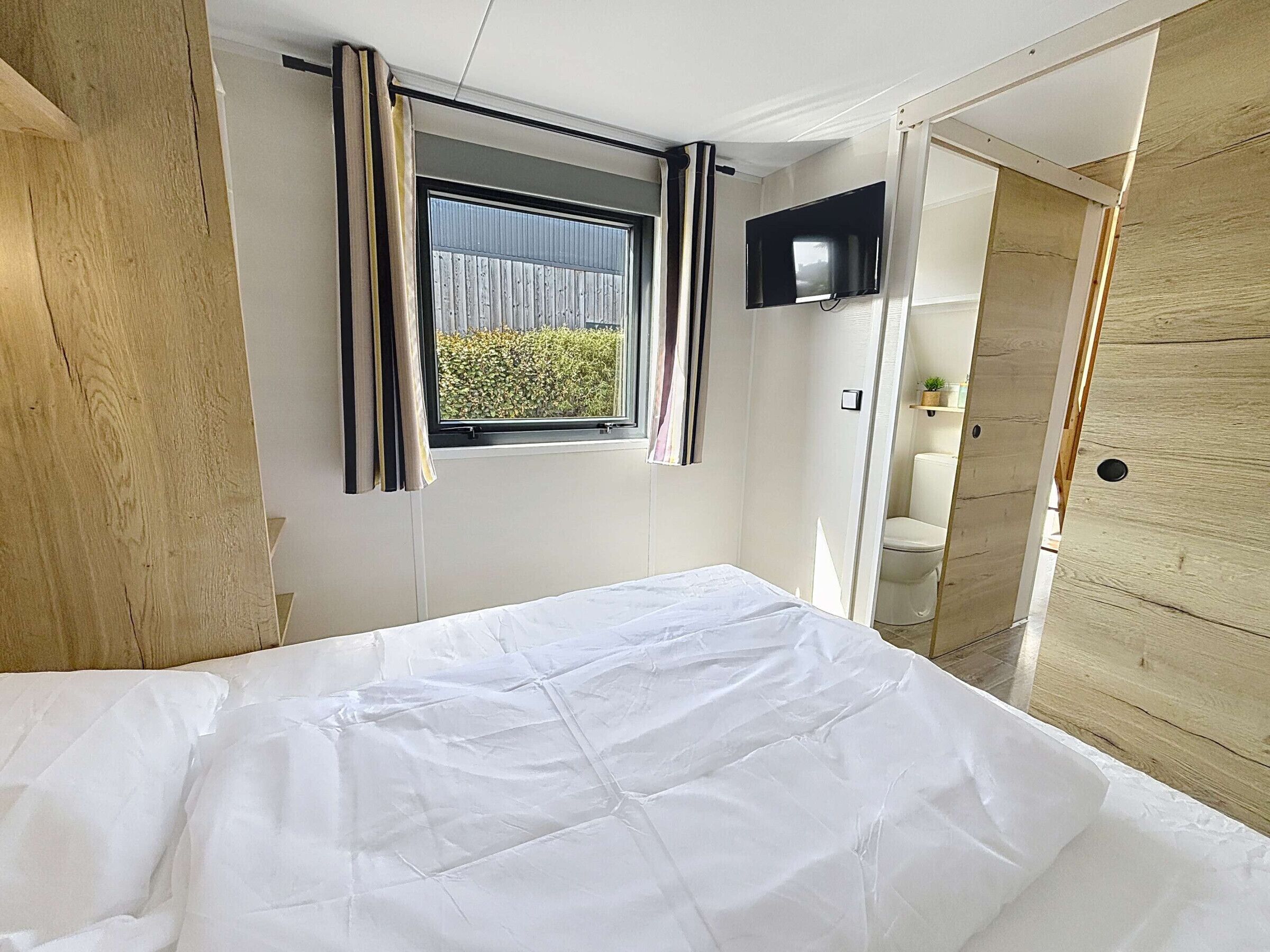 A bedroom with a bed, a window with curtains, a television, and a wooden door.