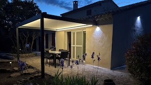 A covered patio area with a table and chairs, surrounded by plants and a stone pathway.