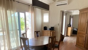 A spacious dining area with a large round table and wooden chairs, a ceiling fan, and a view of the outdoors through large windows with sheer curtains.