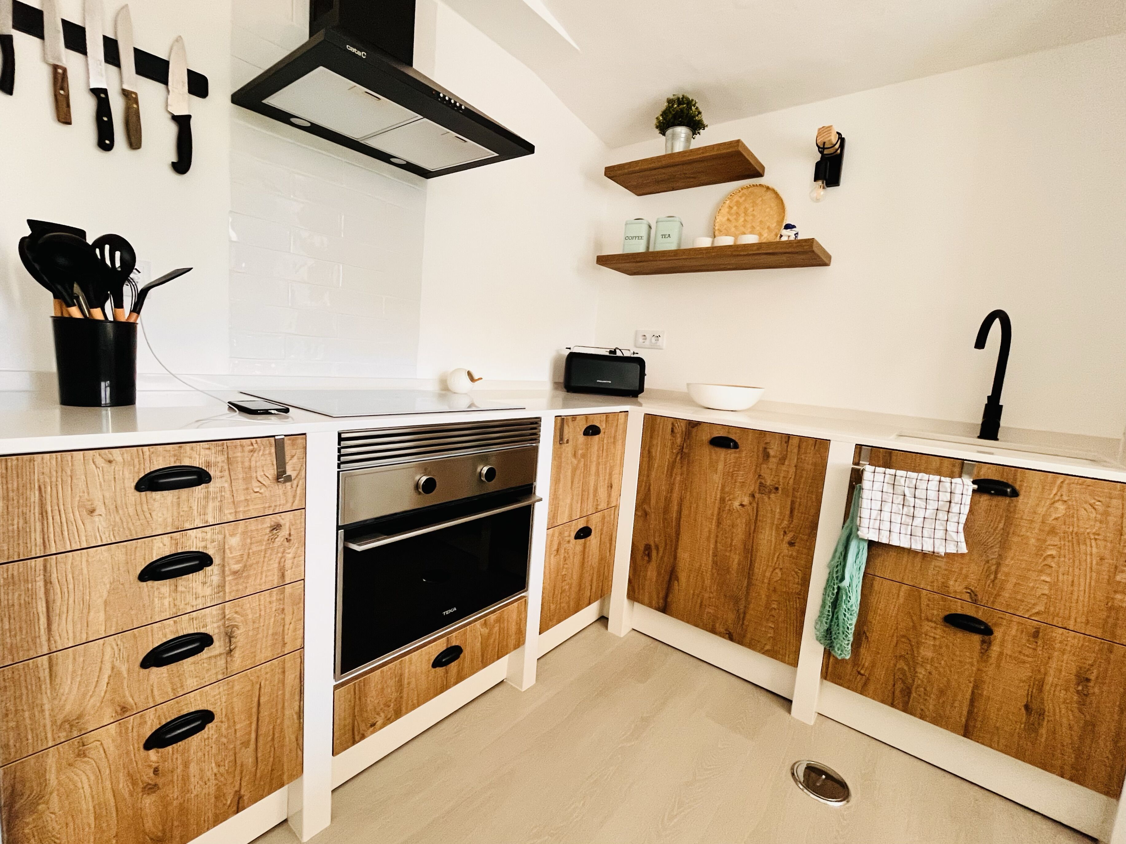 Una cocina moderna con armarios de madera, horno empotrado, campana extractora negra y encimera blanca.
