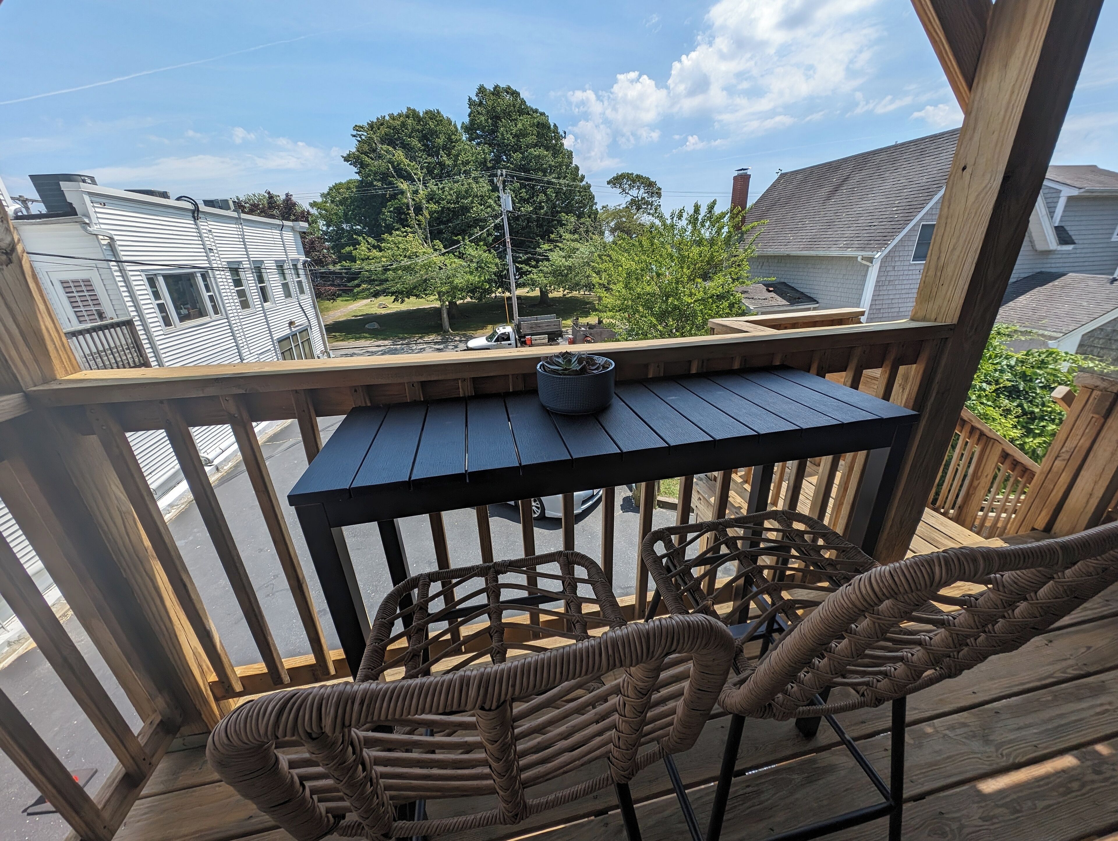 Eine Holzterrasse mit Tisch und Korbstühlen, mit Blick auf ein Wohngebiet mit Häusern und Bäumen.