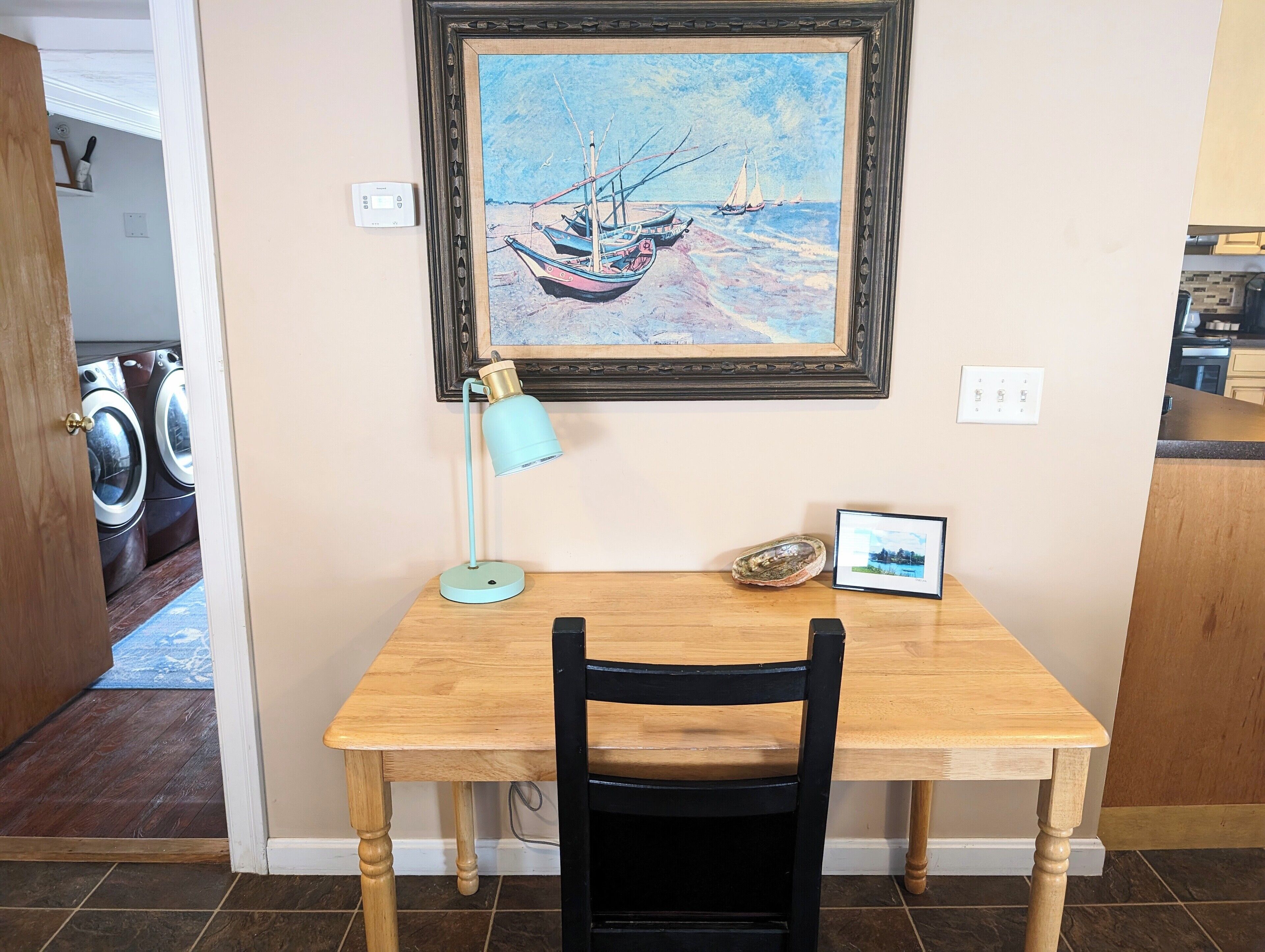 Une pièce avec une table en bois, une chaise noire, un tableau encadré représentant des bateaux, une lampe de bureau bleue et une petite photo encadrée accrochée au mur.