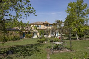 Une maison à deux étages, avec un toit recouvert de tuiles, entourée d’un jardin bien entretenu où se trouvent une table et des chaises.