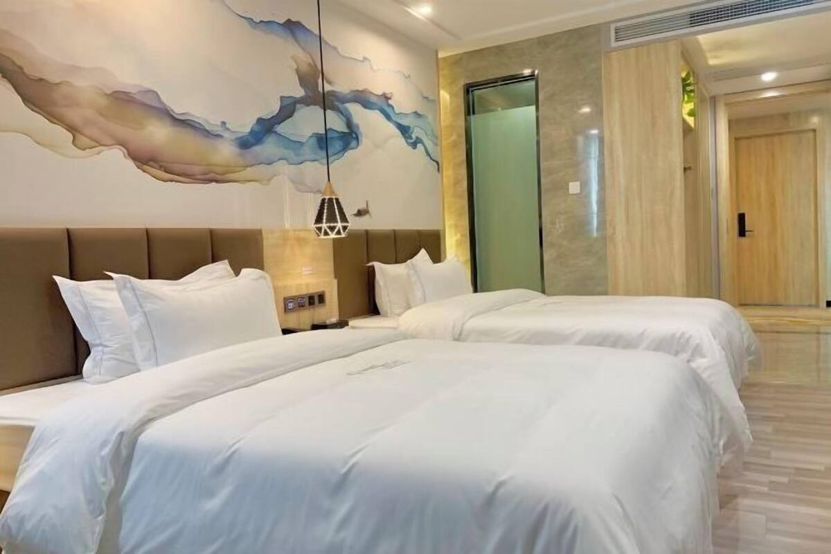 A hotel room with two beds, a large abstract painting, a modern light fixture, and a glass elevator.
