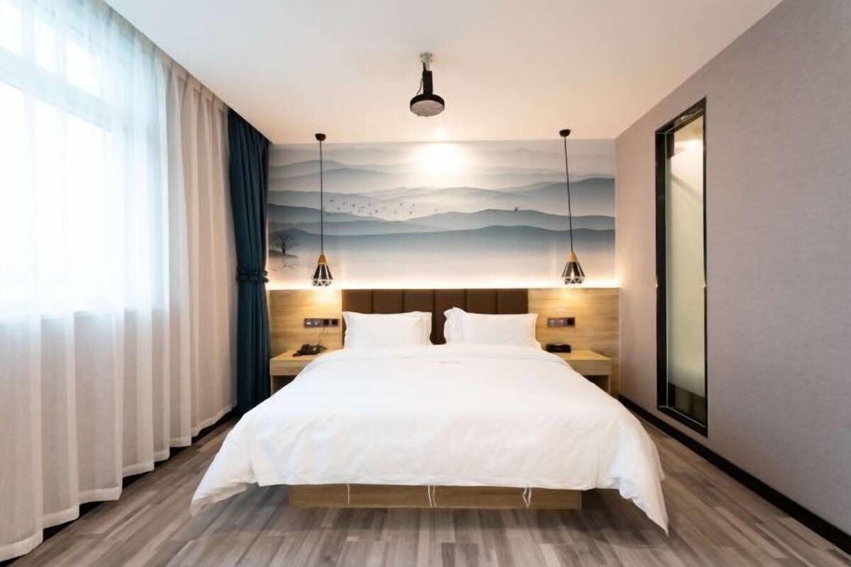A hotel room with a large bed, bedside tables, a nightstand, a wall-mounted lamp, a ceiling light, and a large window with curtains.