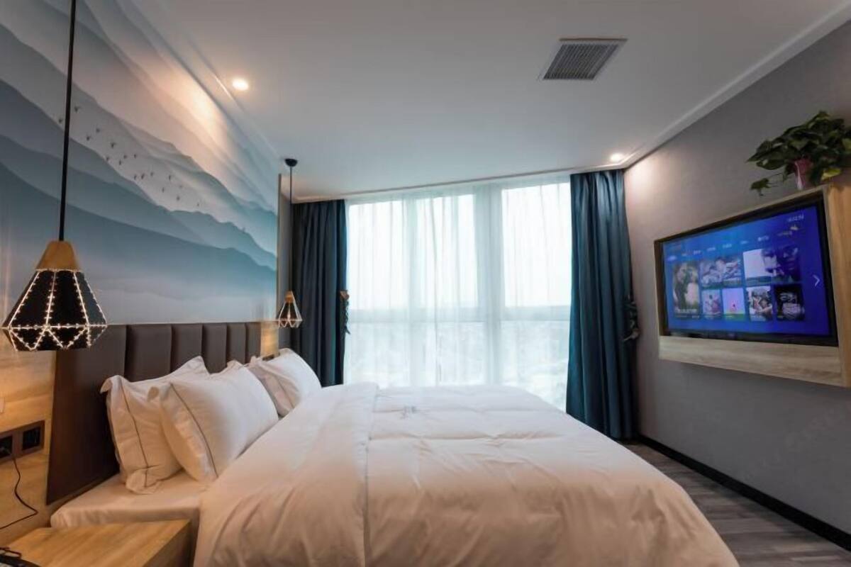 A hotel room with a bed, a TV mounted on the wall, a ceiling mural, and a window with curtains.