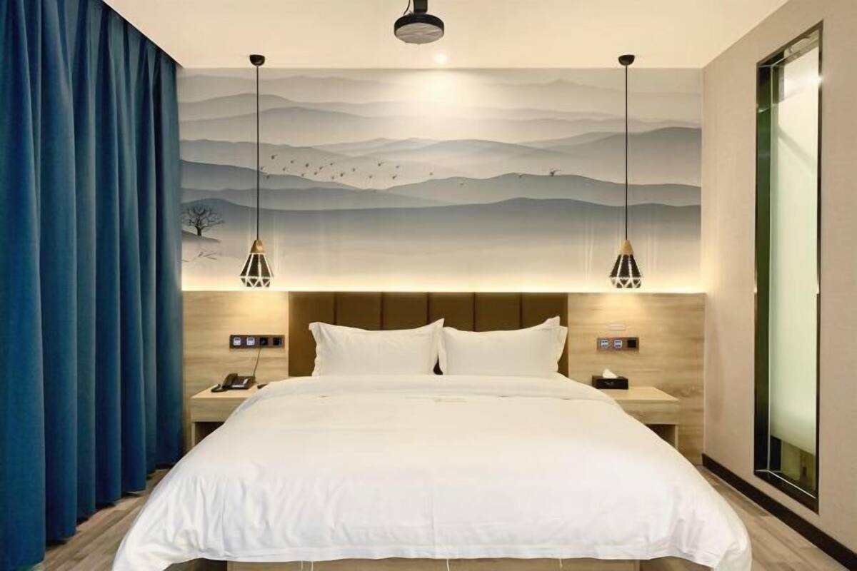 A hotel room with a large bed, two bedside tables, a nightstand, a wall-mounted lamp, and a large window.