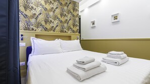 Una habitación de hotel con una cama, almohadas blancas, un cabecero y una pared con estampado floral. Hay dos toallas dobladas sobre la cama.