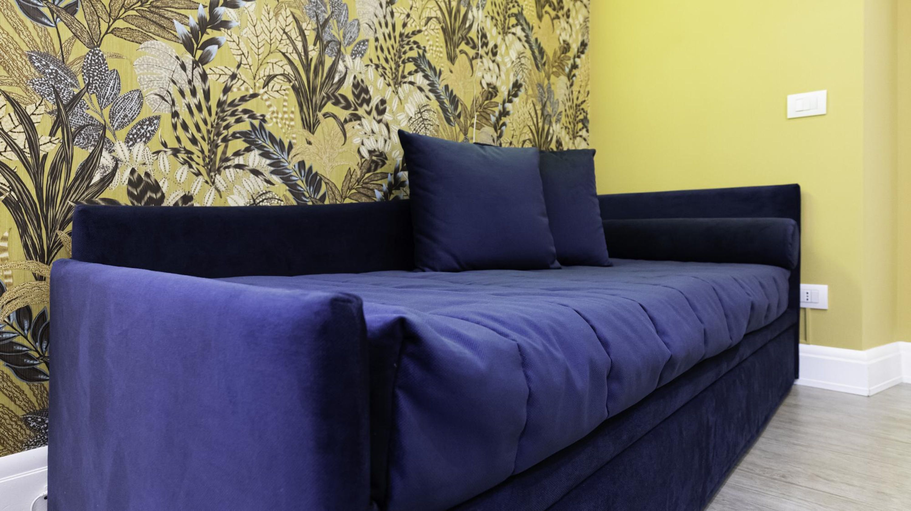 Un sofá azul oscuro con un cojín a juego frente a una pared amarilla con papel tapiz floral.
