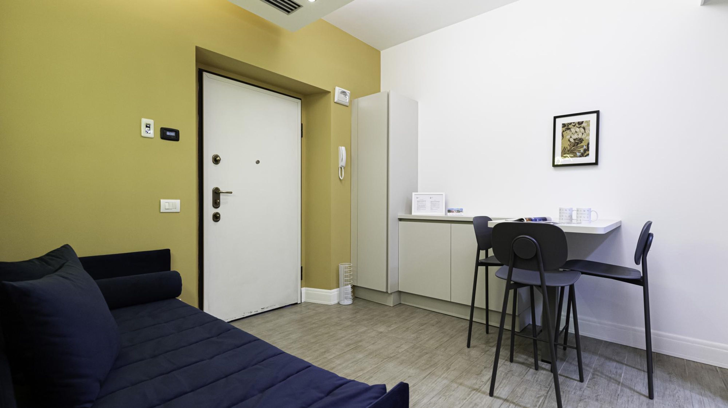 A room with a bed, a desk, and a chair. There is a door with a phone and a light switch on the wall.
