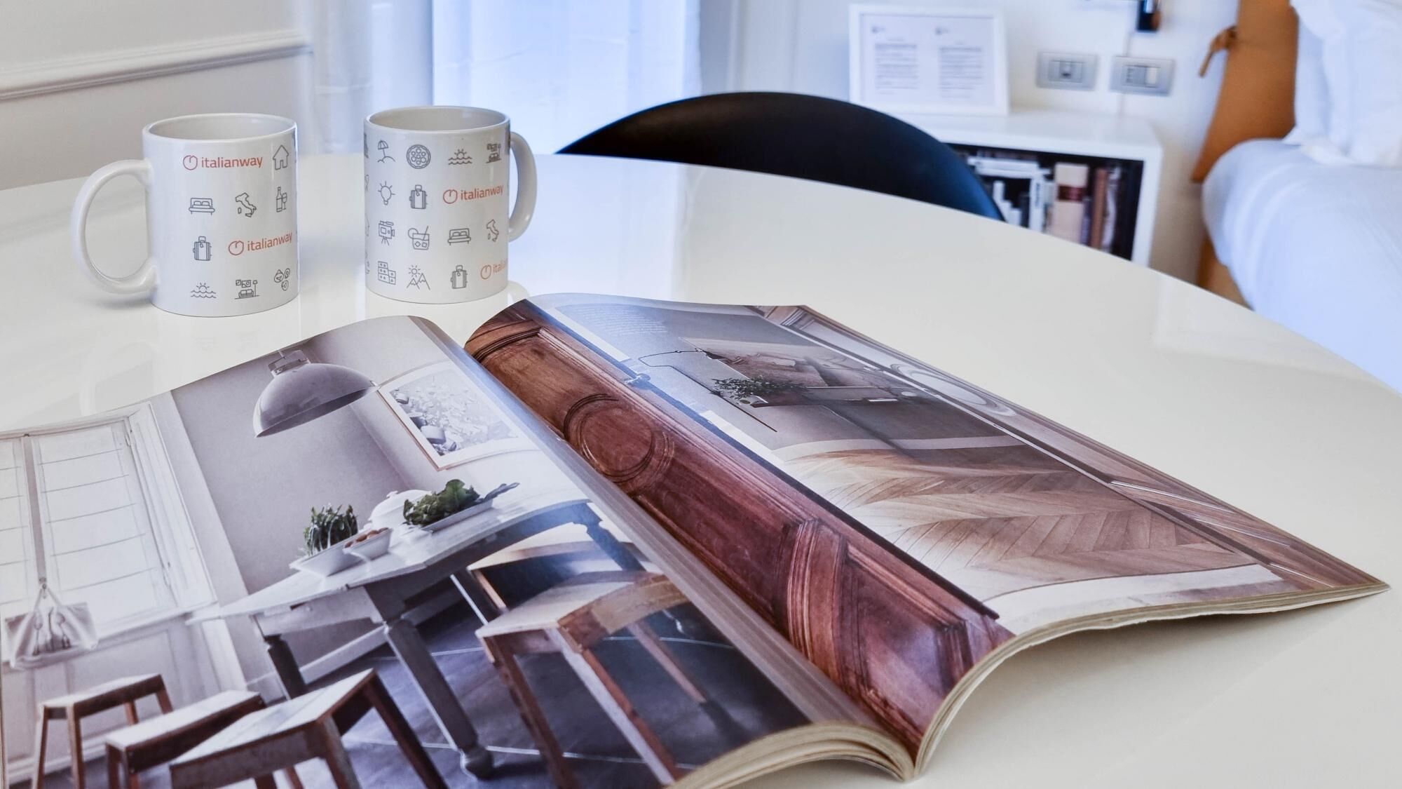 Una mesa con dos tazas blancas que llevan el logotipo de Italianway, una revista con imágenes de arquitectura y una silla de madera al fondo.