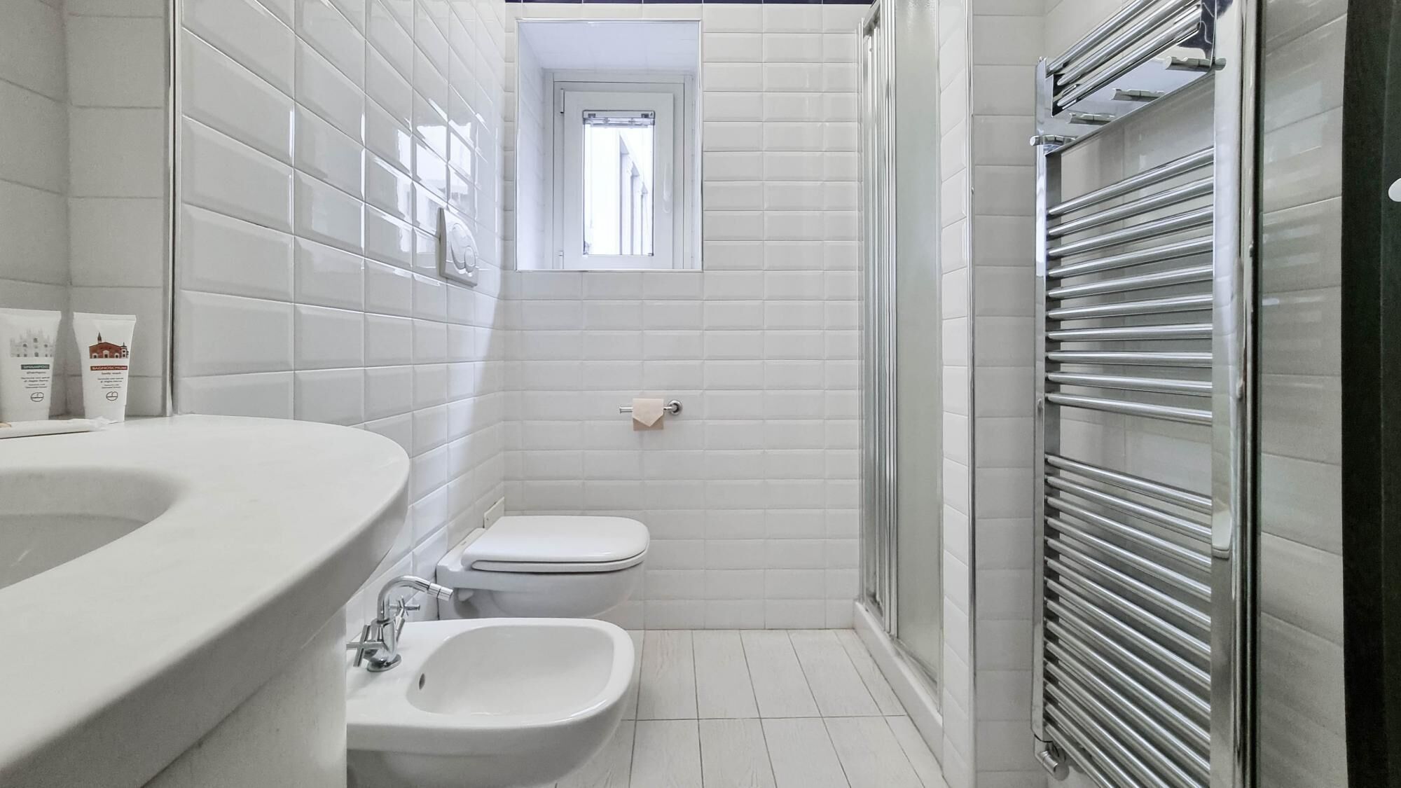 A modern bathroom with white tiles, a bathtub, a toilet, a bidet, and a towel rack.