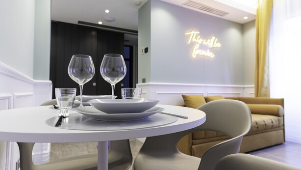 Una mesa de comedor con platos blancos, dos copas de vino y cubiertos. Un letrero de neón en la pared dice 'This will be forever'.