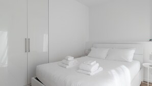 Une chambre à coucher minimaliste, aux murs blancs, avec un lit, des oreillers et des serviettes soigneusement pliées.