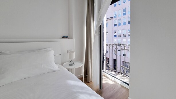 Une chambre d’hôtel avec un grand lit, une petite table de chevet avec une lampe, et une vue sur un immeuble à travers une fenêtre avec des rideaux.