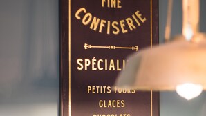 Un tablero de menú con el texto 'Pâtisserie Fine Confiserie' y una lista de productos que incluye 'Petits Fours', 'Glaces', 'Chocolats', 'Marrons Glacés' y 'Macarons'.