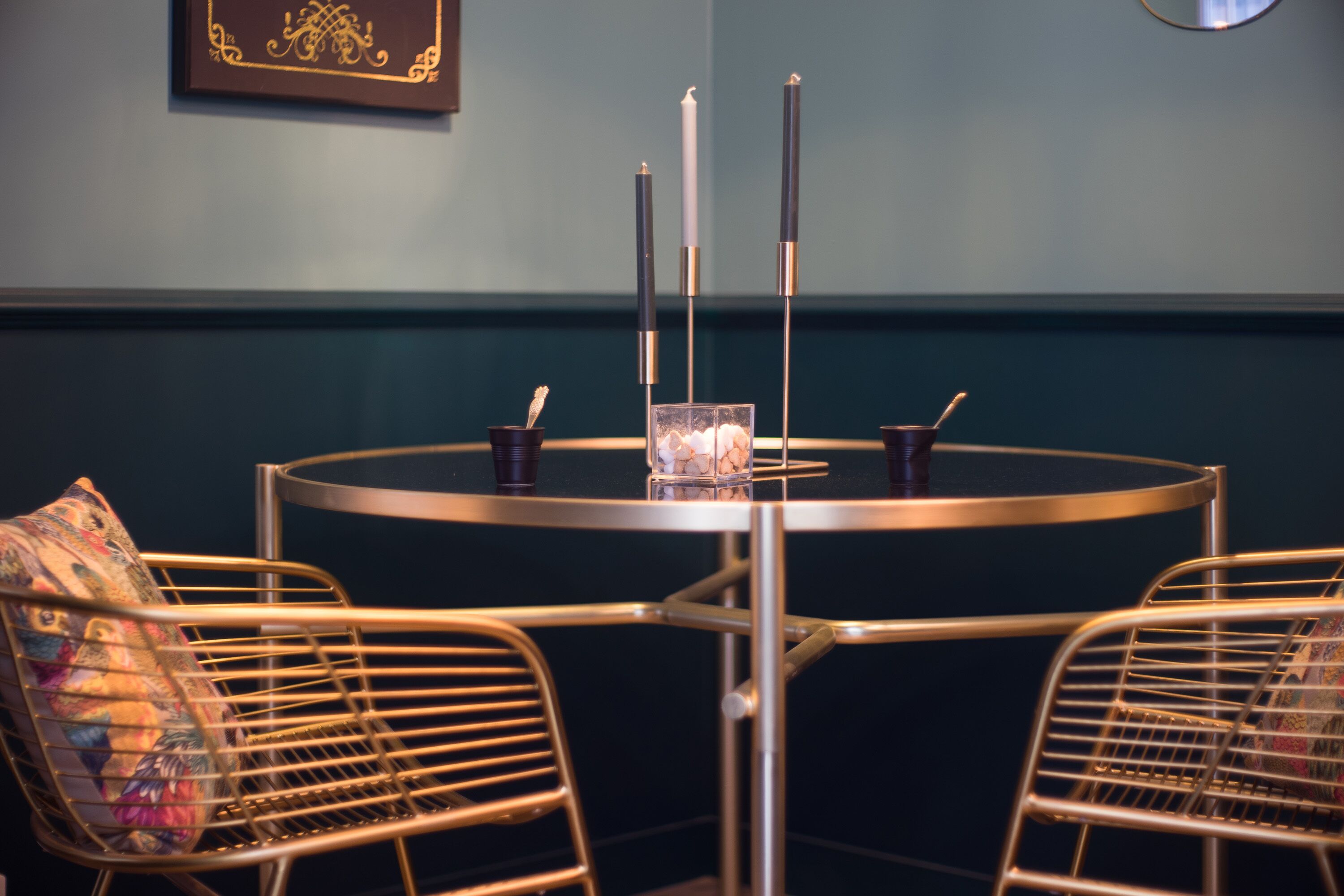 Een ronde tafel met drie kaarsen, een glas met een bloem en een fotolijst. Er zijn twee metalen stoelen met gestreepte kussens.