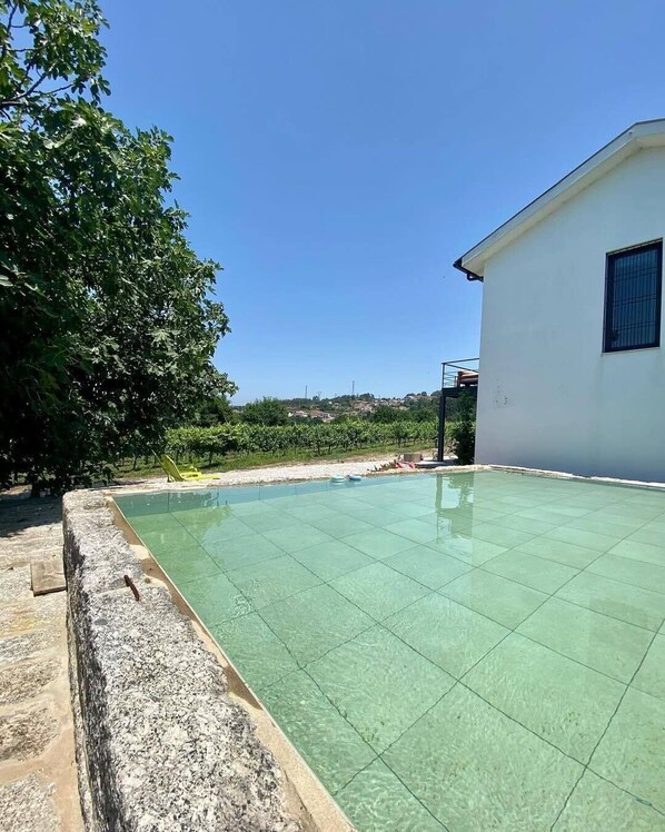 Una piscina con borde de piedra, un edificio blanco con una ventana y un árbol al fondo.