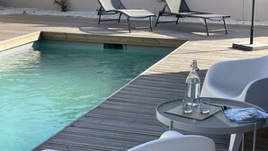 Área ao lado da piscina com deck de madeira, espreguiçadeiras e uma mesa com garrafa e copos.