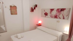 En snyggt bäddad säng med vita lakan och handdukar, en sänglampa, en vägghylla med böcker, ett inramat konstverk med blommotiv och en spegel på väggen.