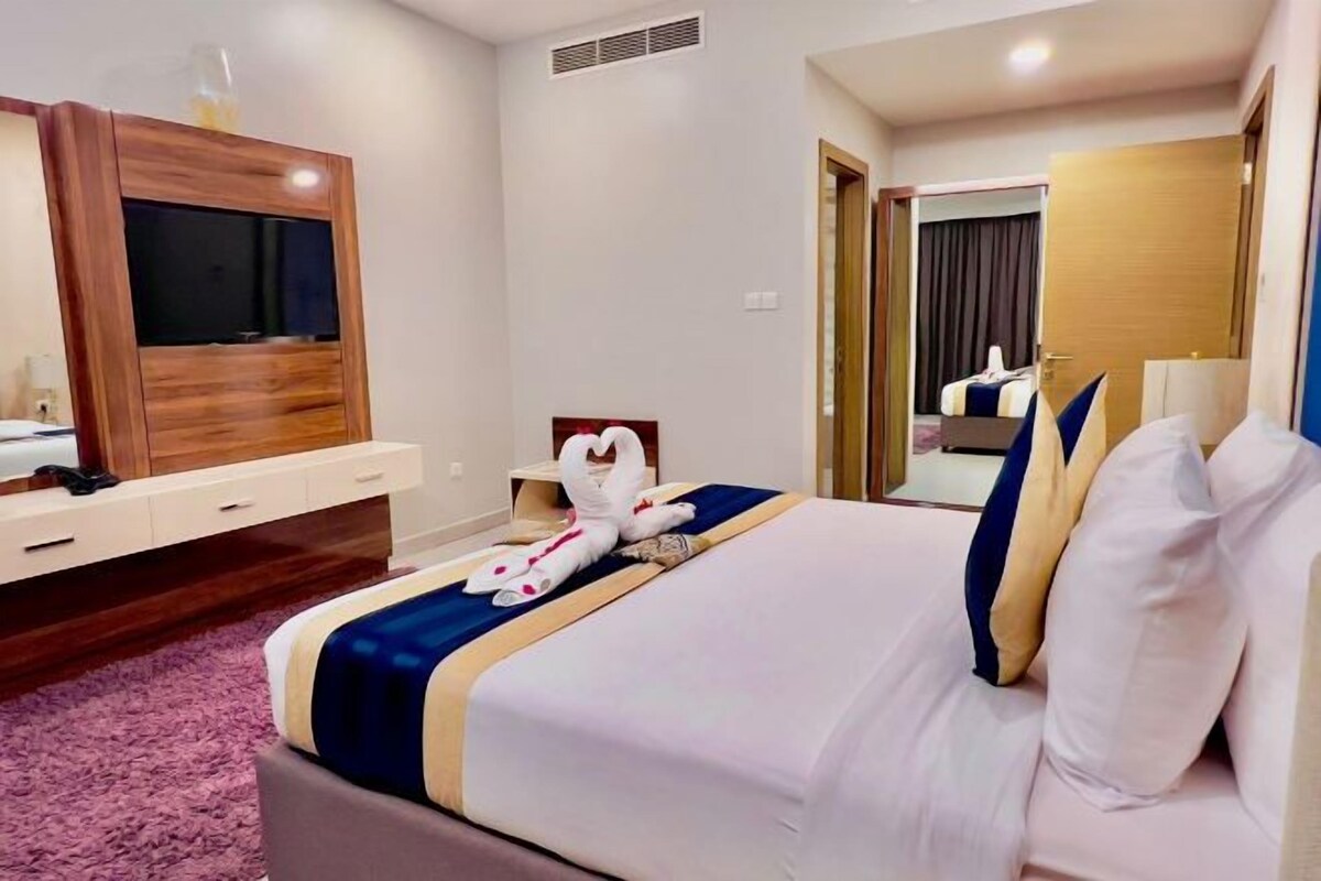 Une chambre d’hôtel avec un grand lit, un téléviseur posé sur une commode en bois, un miroir et une porte donnant sur une autre pièce.