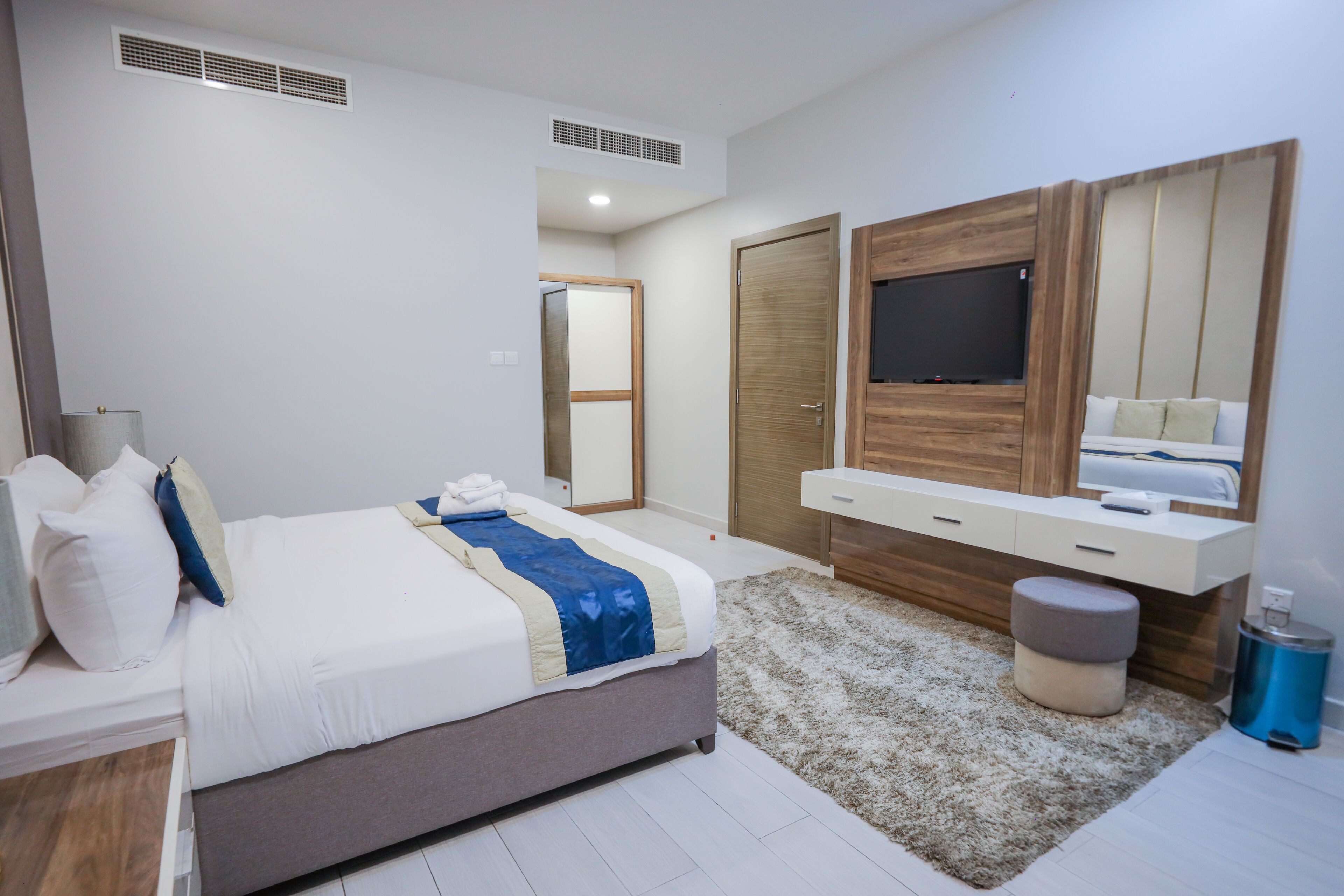 A modern bedroom with a bed, a TV mounted on a wooden panel, a bedside table, and a mirror.