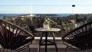 Varanda com mesa redonda e duas cadeiras de vime, com vista para a paisagem costeira com falésias e vegetação.
