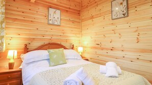 A wooden-paneled room with a bed, two wall-mounted lamps, and two framed floral artworks. - Buttercup Lodge (Leiston)