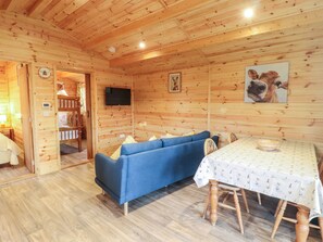 A wooden cabin interior with a blue sofa, a dining table with a patterned tablecloth, and a mounted television. - Buttercup Lodge (Leiston)