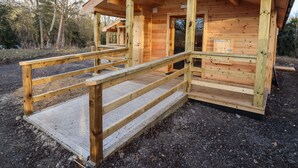 A wooden cabin with a ramp leading to the entrance, surrounded by trees. - Buttercup Lodge (Leiston)