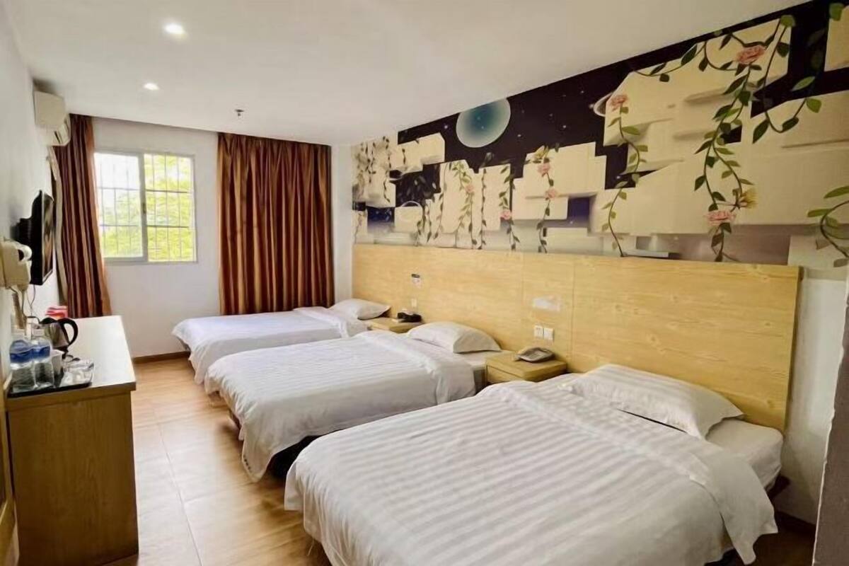 Ein Hotelzimmer mit drei Betten, einem hölzernen Kopfteil, einem Schreibtisch mit Lampe und einem Fenster mit Vorhängen.