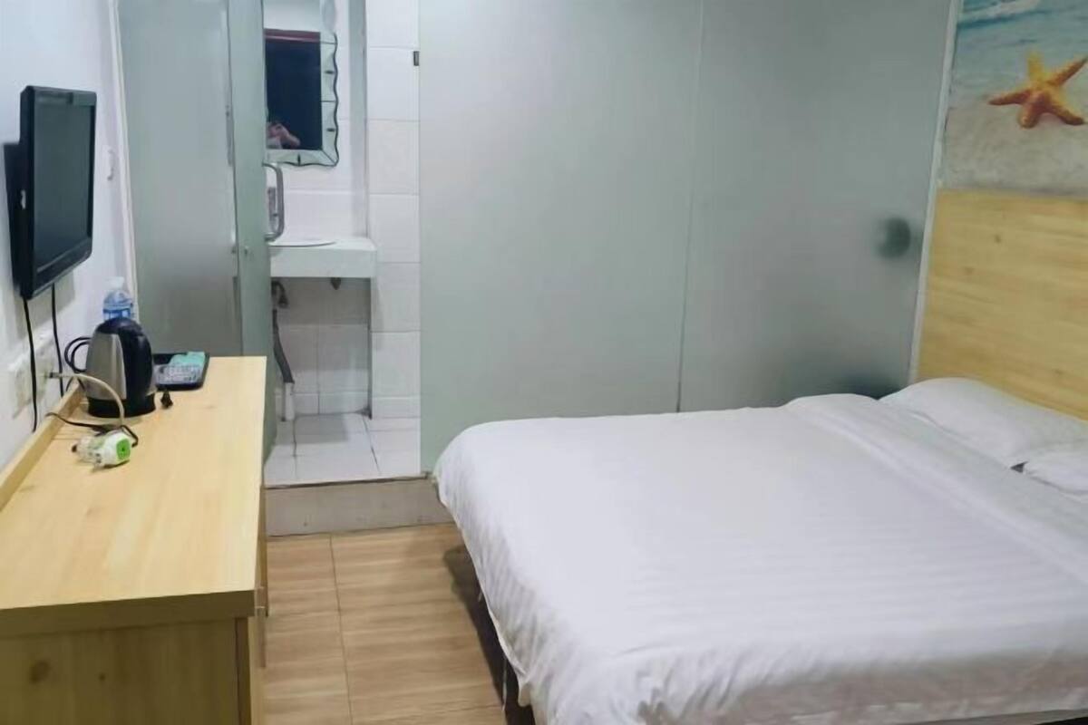 Ein Hotelzimmer mit einem Holztisch, einem Bett mit weißer Bettwäsche, einem an der Wand befestigten Fernseher und einem durchgehenden Türrahmen einsehbaren Badezimmer.