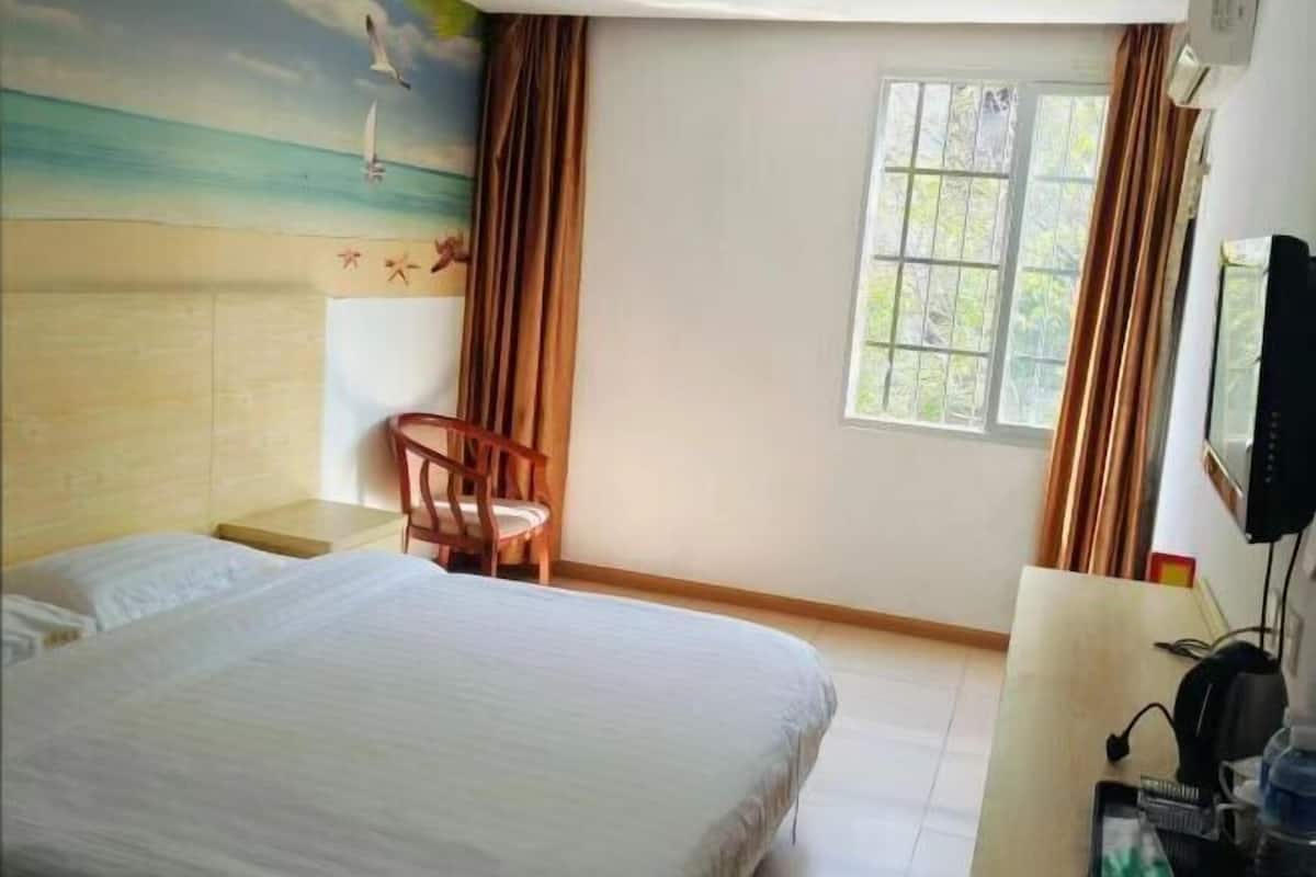 Ein Hotelzimmer mit Bett, Stuhl, Schreibtisch, Fenster und Wandgemälde mit Strandmotiv.