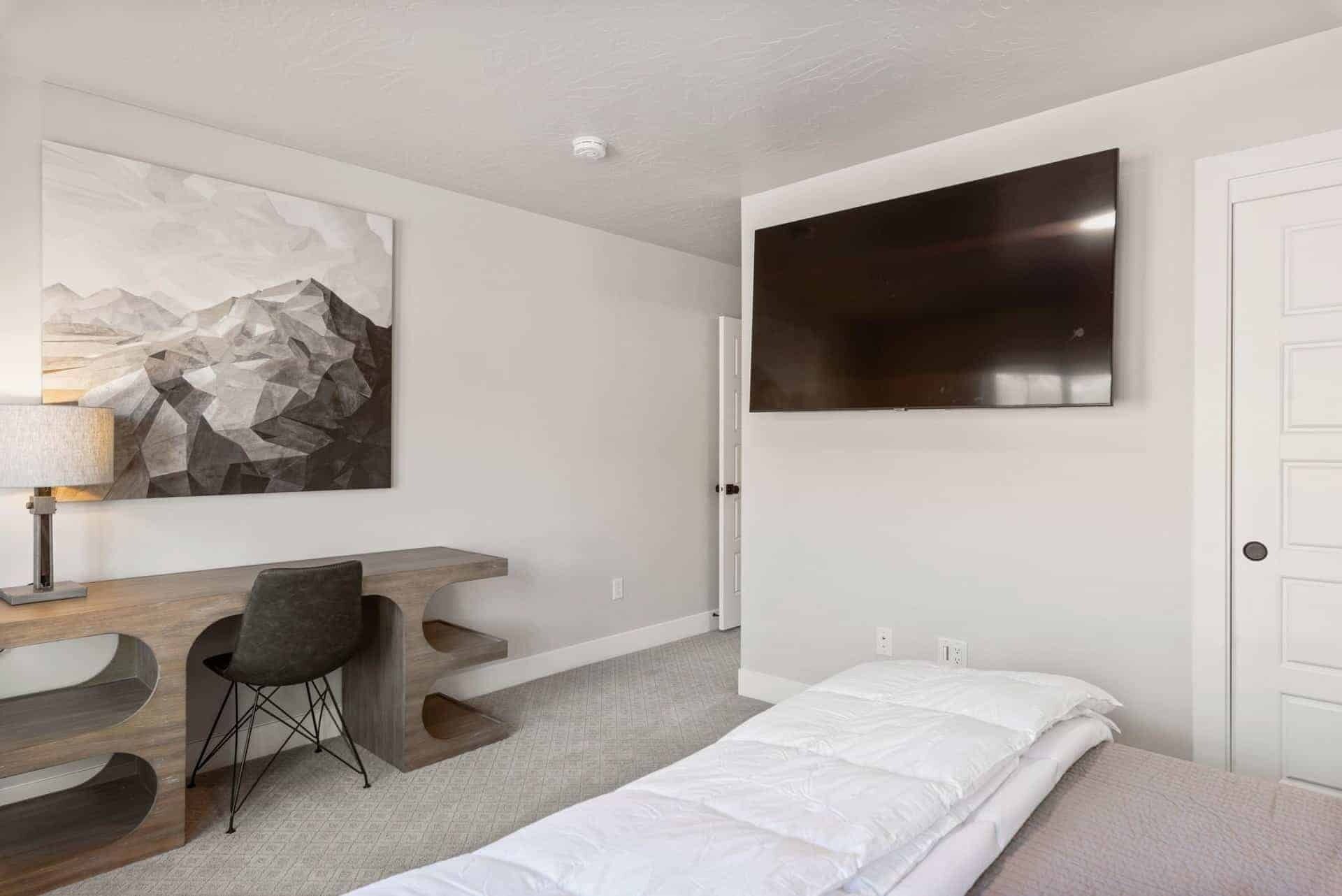 A modern bedroom with a bed, a desk, a chair, a TV, and a mountain artwork on the wall.