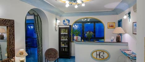 A reception area with a blue-tiled floor, a reception desk with a logo, a chandelier, and a view through a window.