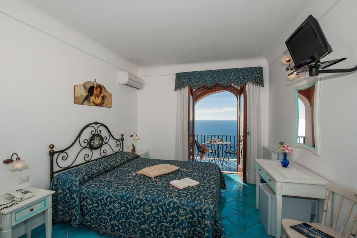 A bedroom with a bed, a desk, a chair, a TV, and a view of the sea.