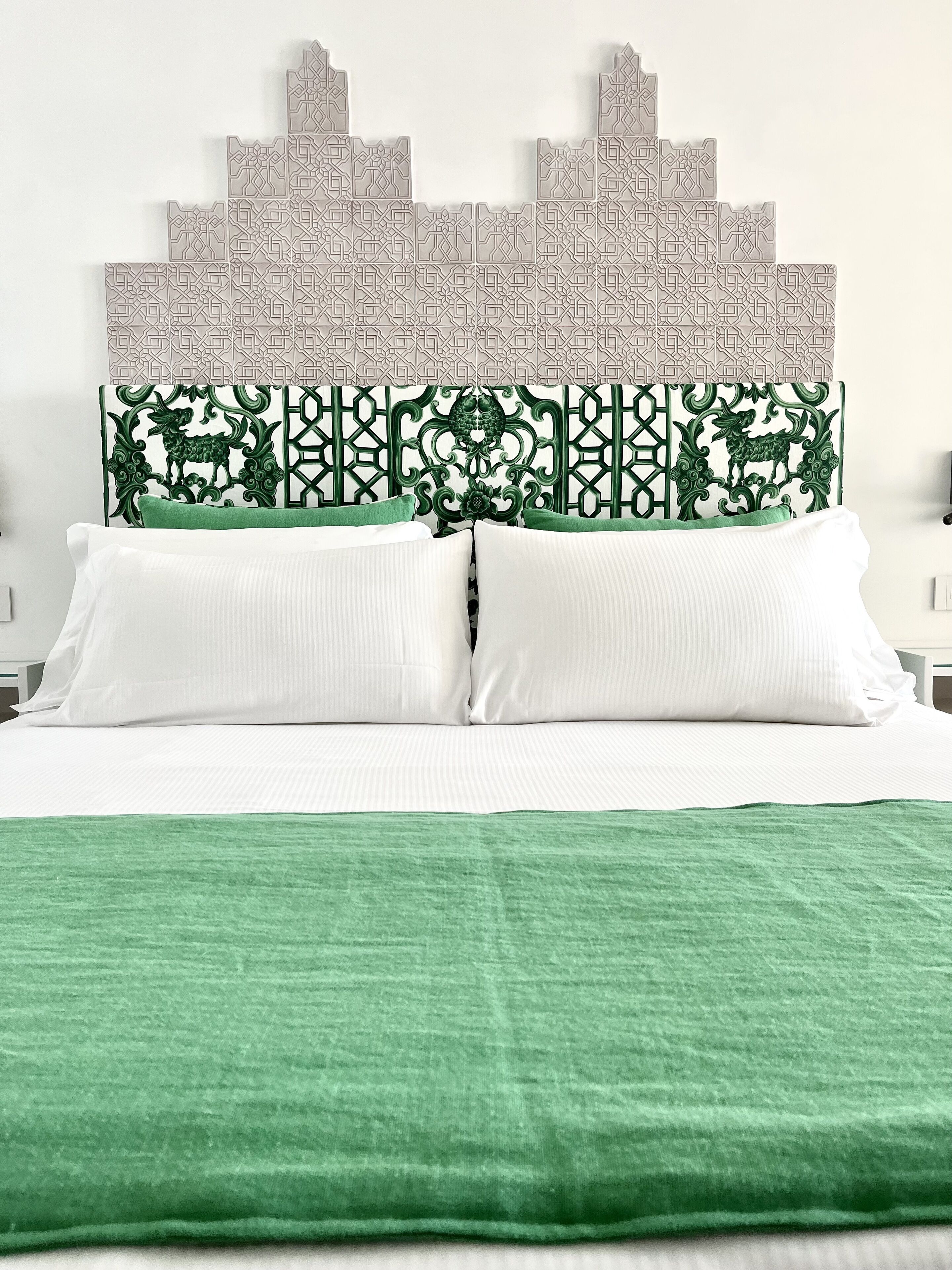 Una cama con sábanas blancas, un plaid verde y un cabecero decorativo con un estampado geométrico.