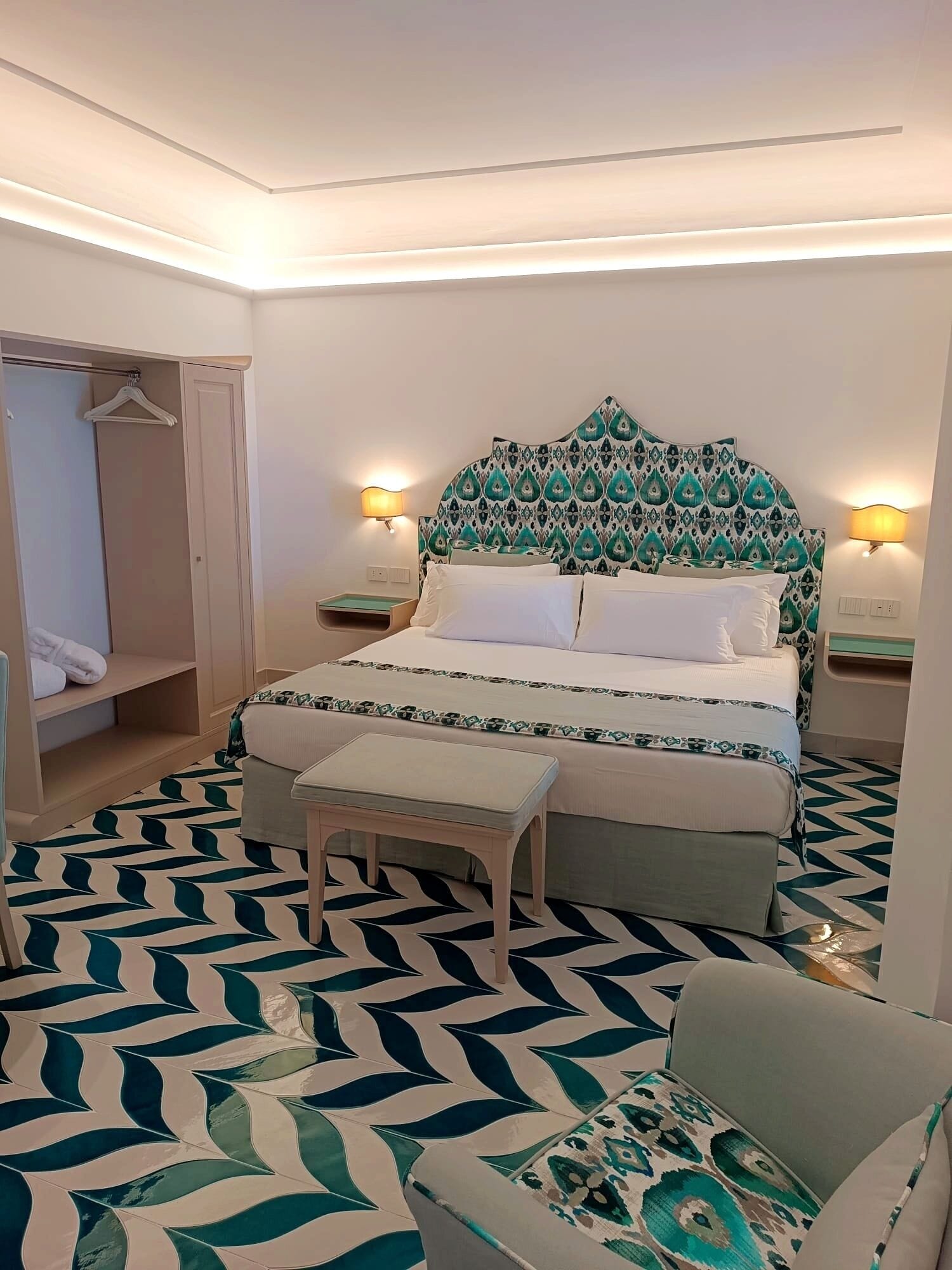 A hotel room with a bed, bedside tables, a bench, a wardrobe, and a patterned floor.