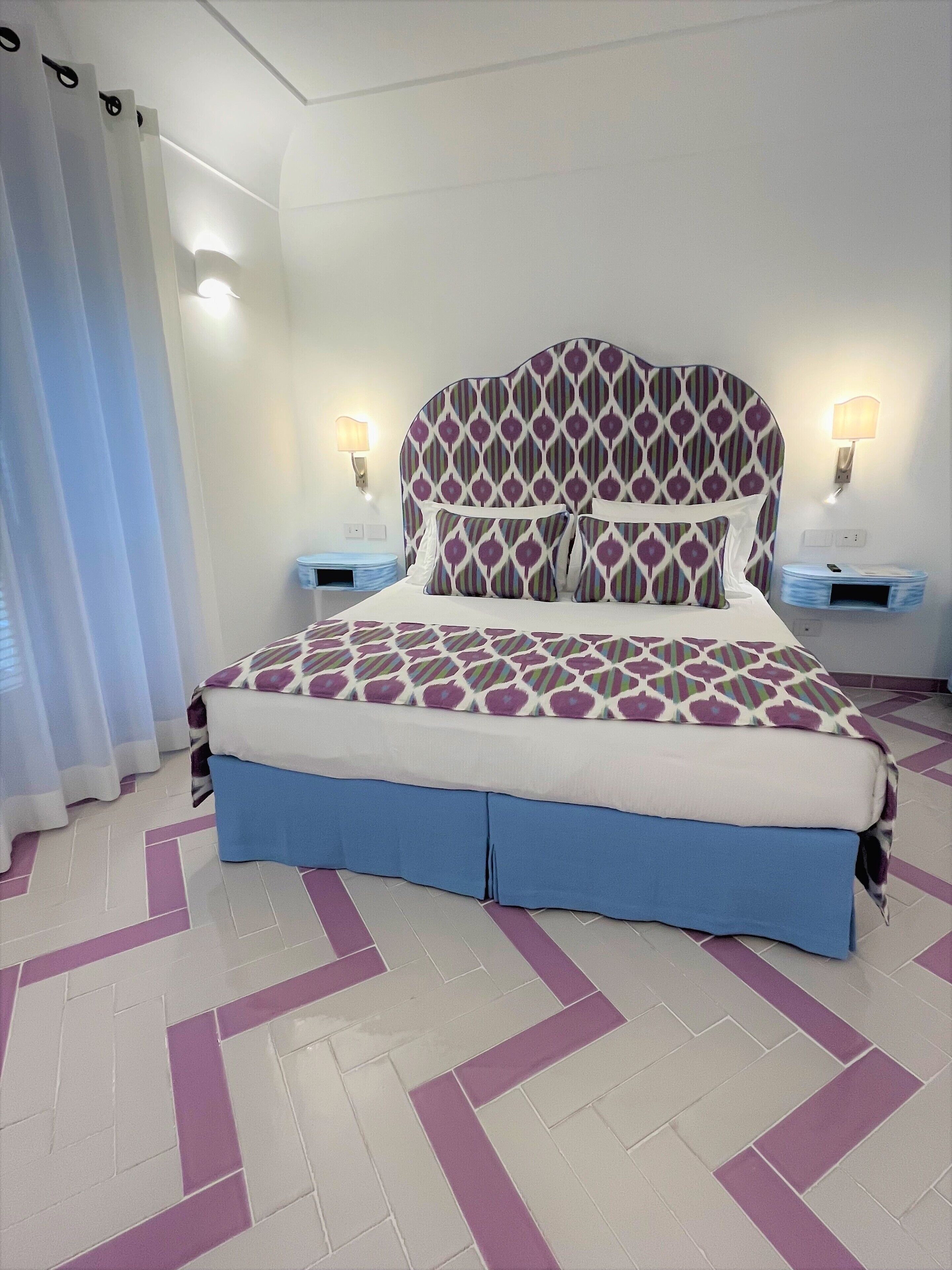 A neatly made bed with patterned bedding, a headboard with a geometric design, two bedside tables with lamps, and a chevron-patterned floor.