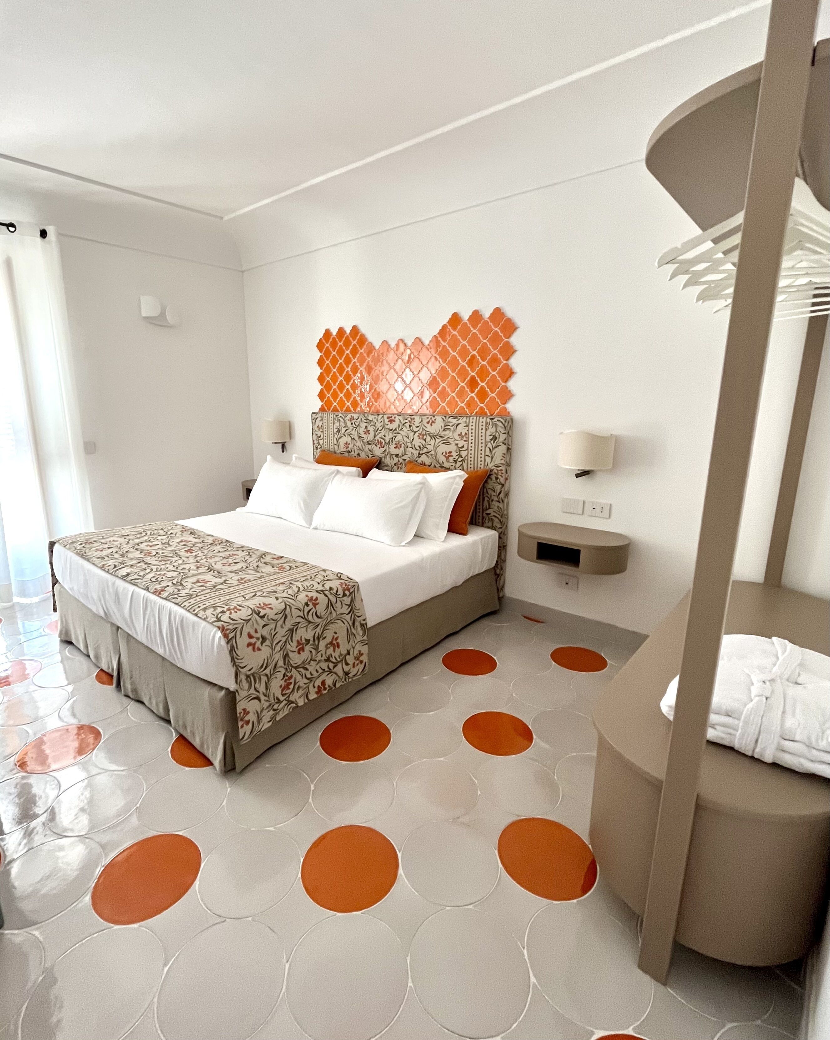 A modern hotel room with a bed, a headboard with a decorative panel, a bedside table with a lamp, and a circular seating area with a towel.