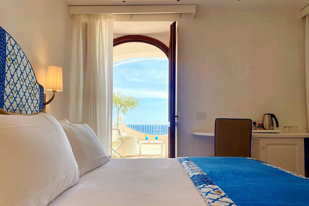 A hotel room with a bed, a desk with a kettle, and a view of the sea through an open door.