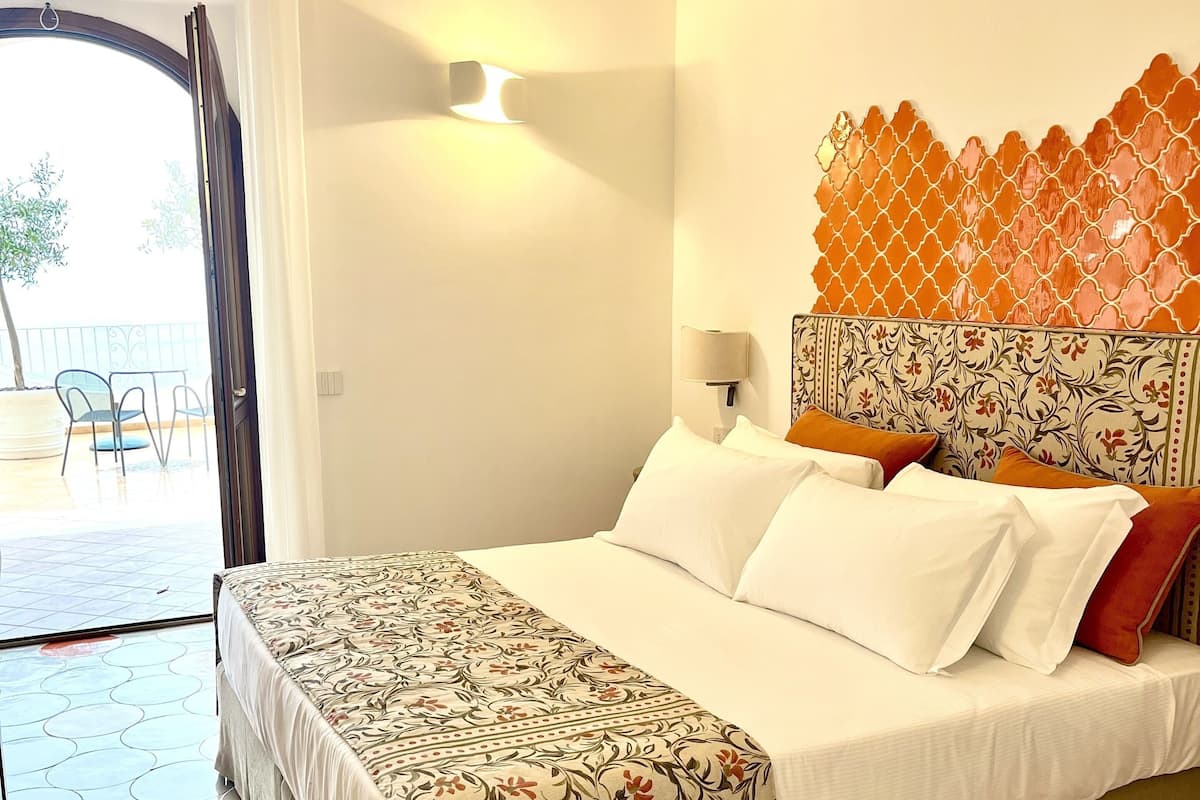 A hotel room with a bed, a patterned headboard, a nightstand, a lamp, and a view of the outside.