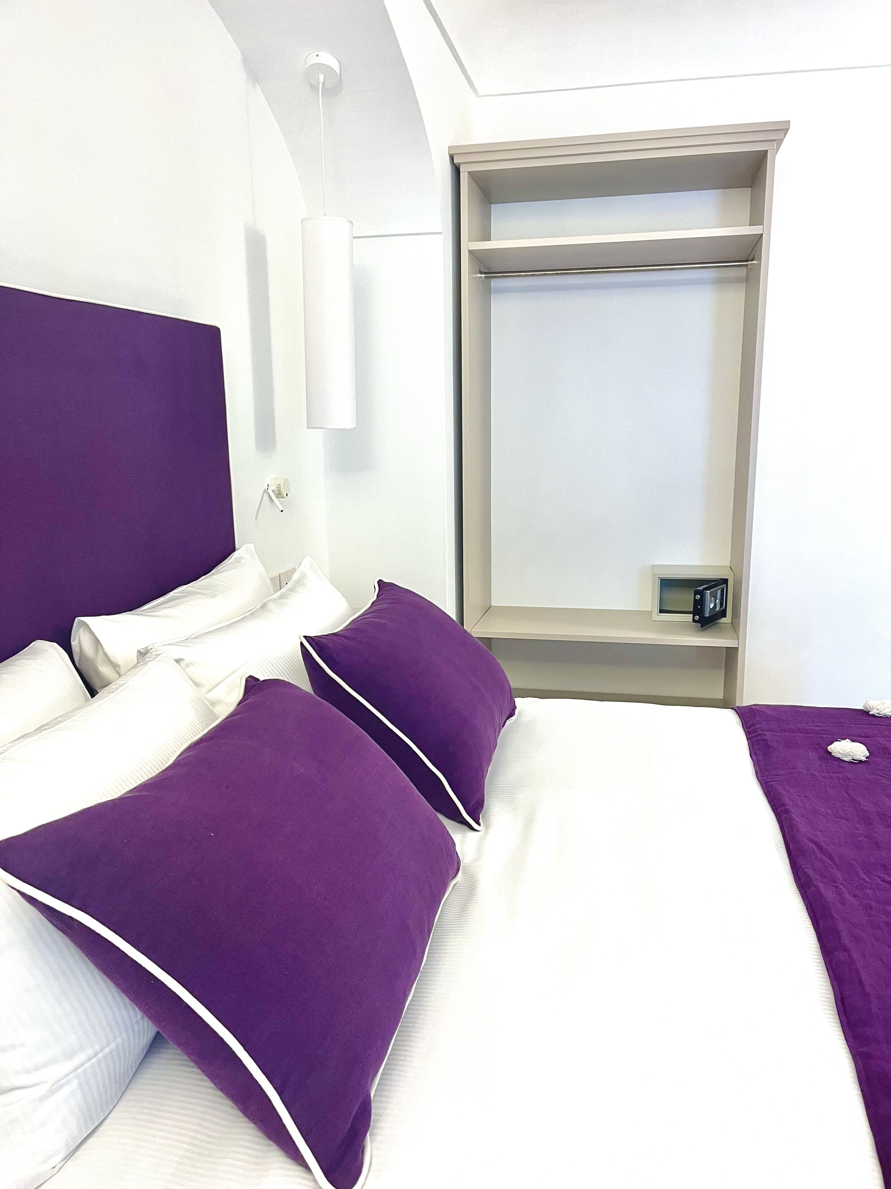 A hotel room with a bed, two purple pillows, a white headboard, a bedside table with a clock, and a window with a view.